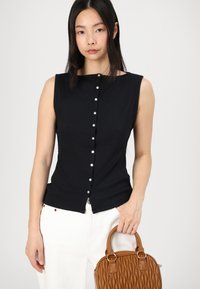 Haut sans manches noir avec une texture côtelée et un décolleté arrondi, comportant une rangée de boutons en métal. Le modèle tient un sac à main matelassé marron.