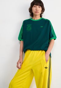 Green cotton t-shirt with black and green "Adidas Originals" logo, paired with yellow polyester pants featuring black stripes and logo.