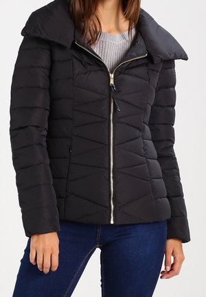 Femme portant une doudoune matelassée noire avec un col haut, une fermeture éclair sur le devant, un pull gris et un jean bleu foncé.