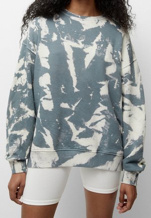 Femme portant un sweat-shirt tie-dye bleu et blanc oversize avec le texte "A TRIP ABROAD" et un short cycliste blanc, debout devant un fond uni.