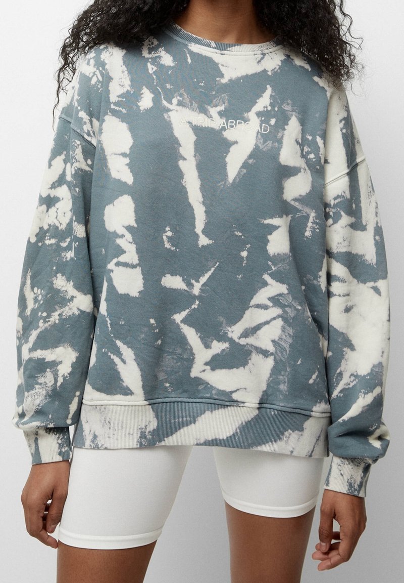 Femme portant un sweat-shirt tie-dye bleu et blanc oversize avec le texte "A TRIP ABROAD" et un short cycliste blanc, debout devant un fond uni.