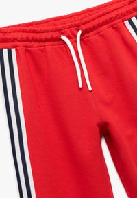 Røde sweatpants med elastisk talje, hvid snøre og sort-hvide sidelister. Blødt, struktureret stof er synligt.