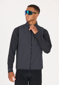 Svart lättvikts cykeljacka med hög krage och full dragkedja. Har lång ärmar och en texturerad design. Bärs med stora reflektiva solglasögon.