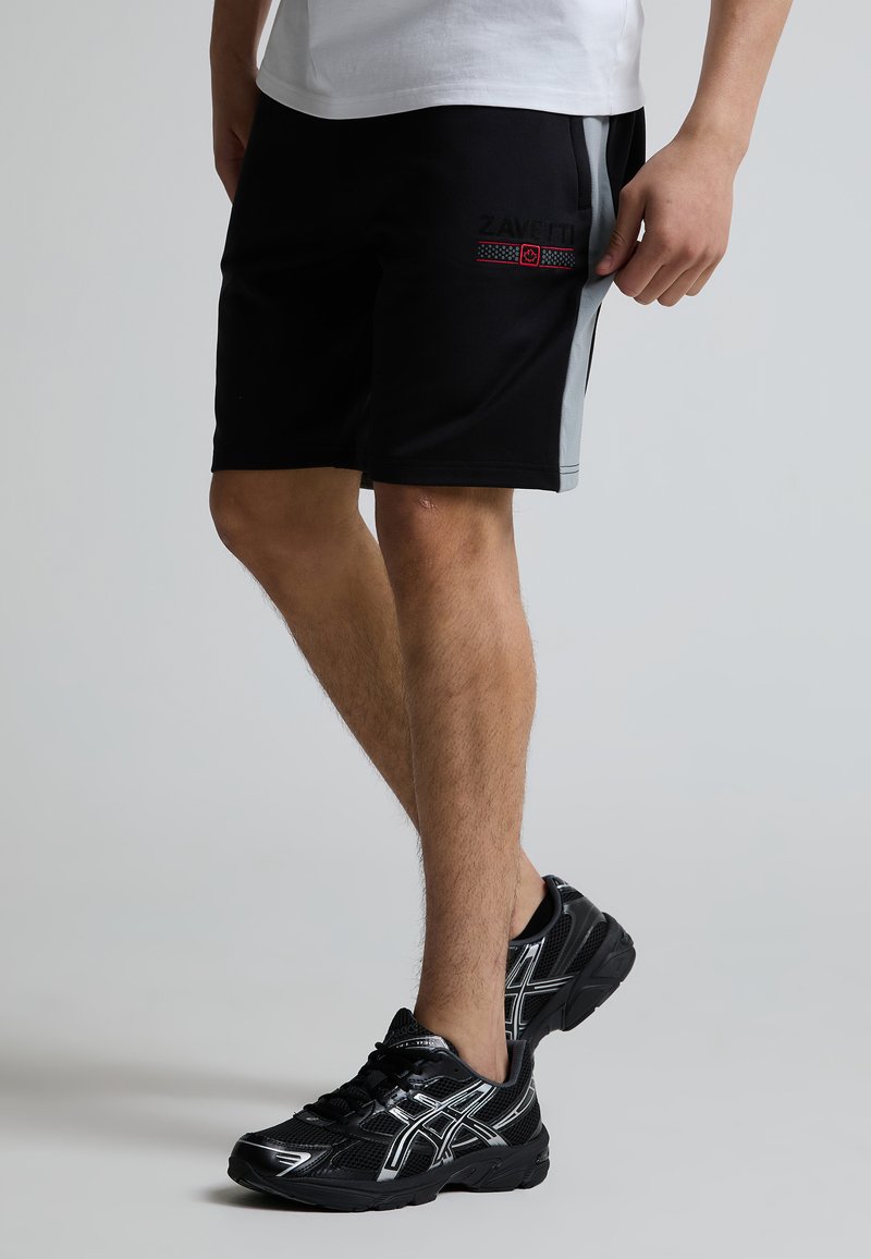 Homme portant un short de sport noir avec une bande latérale grise et des chaussures de course noires sur fond gris clair.