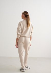 Completo loungewear beige in fleece con top dalla vestibilità ampia e pantaloni tapered, abbinato a sneaker grigie e argentate. Tessuto morbido, design rilassato.