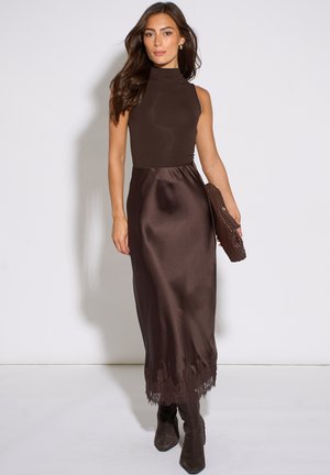 Woman wearing sleeveless brown top and satin midi skirt with lace trim, holding woven clutch and wearing brown boots, posing against white background.