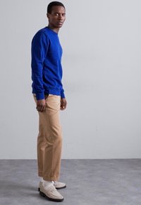 Modra crewneck svetilka, peščene prilagojene hlače, bele nogavice in loaferji. Gladek material, prilegajoča silhueta, sproščen stil. Svetlo siva podlaga.