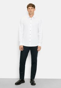 White long-sleeve button-up shirt with a pointed collar, dark buttons, paired with navy trousers and black shoes. Smooth fabric texture.