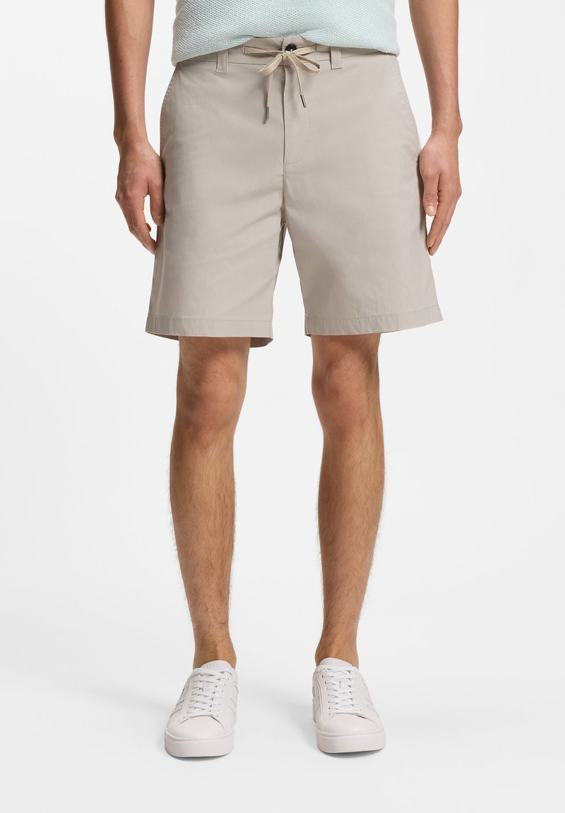 Homme portant un short beige mi-cuisses avec taille à cordon et des baskets blanches, debout sur un fond blanc uni.