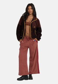 Bordowa i czarna polarowa kurtka z żółtymi wykończeniami, zestawiona z szerokimi spodniami w paski w kolorach czerwonym i brązowym. Czarne botki za kostkę dopełniają całość.