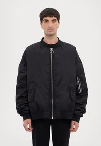 Giubbotto bomber nero in nylon con zip frontale, due tasche laterali e una tasca sul braccio con zip. Presenta polsini e colletto a coste.