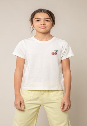 Jeune fille debout, portant un t-shirt blanc avec un petit motif de cerise sur la poitrine et un pantalon jaune clair, sur un fond beige.