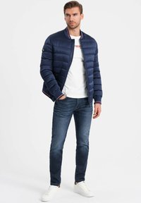 Marineblaue Pufferjacke mit Reißverschluss, gerippten Bündchen und rot-weißen Streifenakzenten, kombiniert mit einem weißen T-Shirt und blauen Jeans. Weiße Sneaker.