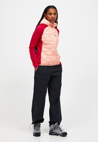 Chaqueta acolchada multicolor con la parte superior en rosa y marrón, que presenta un cierre de cremallera, capucha y pantalones cargo oscuros combinados con botas de senderismo grises.