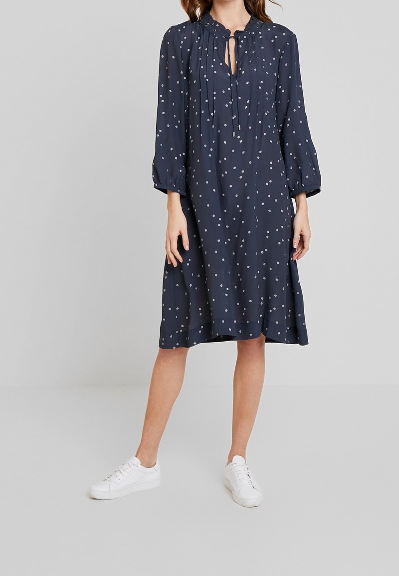 Námořnické modré šaty s dlouhými rukávy, plisovaným živůtkem a malými bílými puntíky. Mají zavazovací krk a volný, splývavý siluetu. Nosit s bílými teniskami.