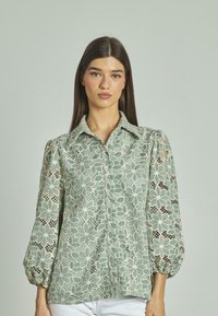 Blusa in pizzo verde chiaro con motivo floreale, colletto strutturato e maniche a sbuffo. Chiusura frontale con bottoni, texture morbida e design a trama aperta.