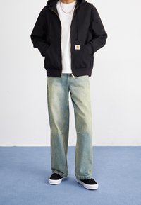 Schwarzer Kapuzenpullover mit Reißverschluss und zwei Vordertaschen, weißes T-Shirt darunter, hellblaue weite Jeans und schwarze Slip-On-Schuhe auf blauem Hintergrund.