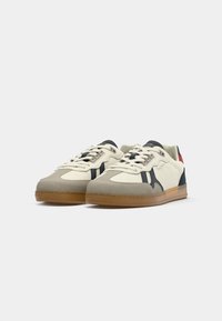 Une paire de baskets basses beige et marine avec des semelles en gomme, des lacets et un subtil logo de la marque sur le côté et la languette.
