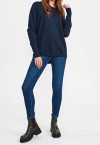 Sudadera azul marino con puños y dobladillo acanalados, combinada con vaqueros ajustados azules y botines verdes con suelas negras. Diseño minimalista, colores sólidos.