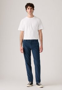 Tânăr stând în picioare, purtând un tricou alb simplu, pantaloni bleumarin și adidași negri cu tălpi albe, pe un fundal deschis și neutr.