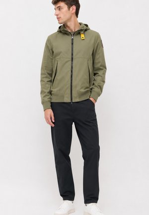 Jeune homme portant une veste à capuche vert olive, un pantalon noir et des baskets blanches, debout avec une main dans la poche, regardant vers la gauche.