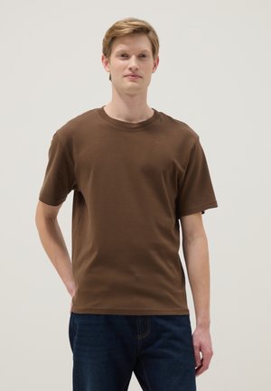 Jonge man die recht vooruit kijkt, gekleed in een bruin T-shirt met korte mouwen en donkerblauwe spijkerbroek, tegen een effen lichte achtergrond.