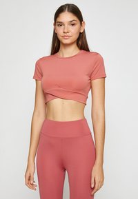 Camiseta corta rosa hecha de material elástico, con cuello redondo y mangas cortas, que presenta un diseño frontal retorcido. Combinada con leggings a juego.