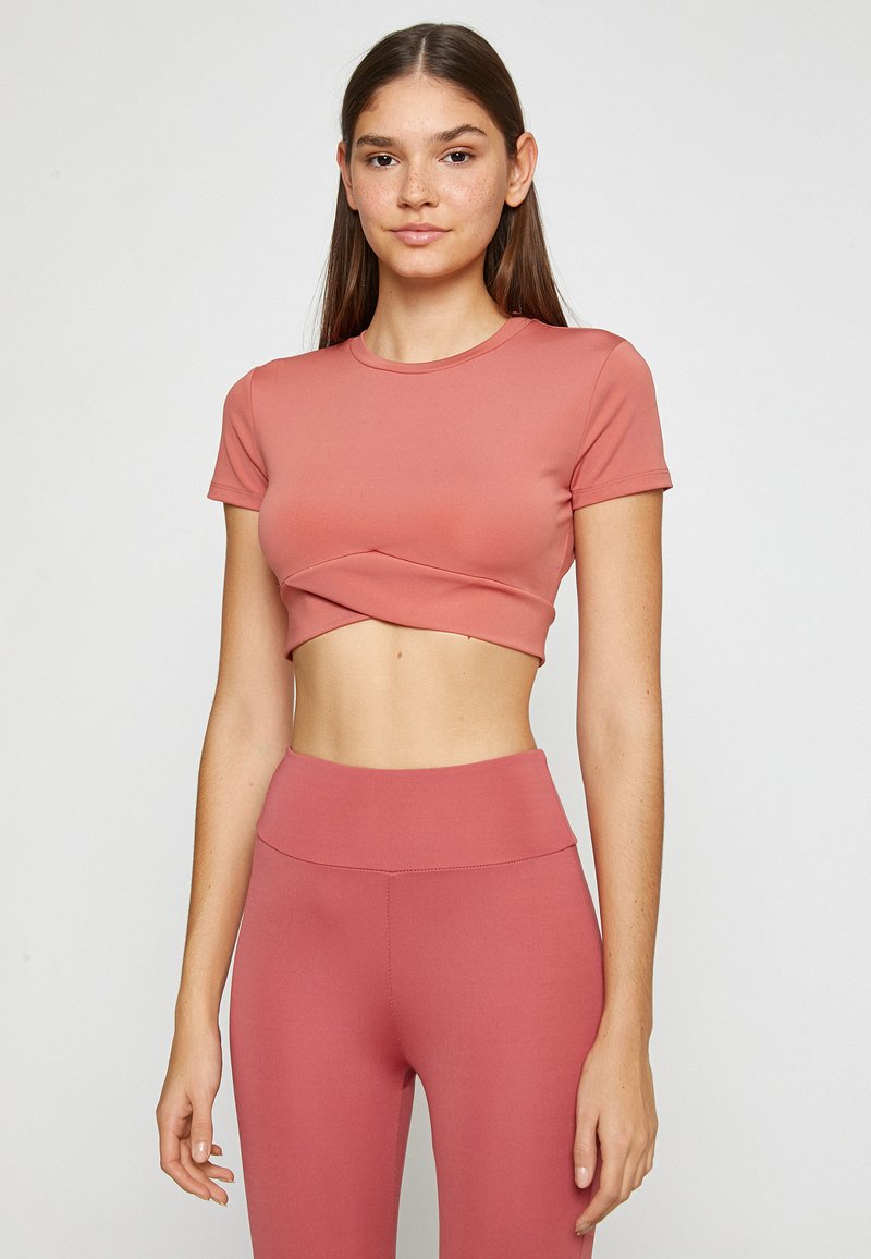 Camiseta corta rosa hecha de material elástico, con cuello redondo y mangas cortas, que presenta un diseño frontal retorcido. Combinada con leggings a juego.