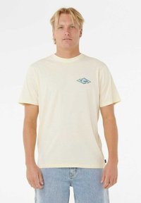 Kortærmet t-shirt i cremefarve lavet af blødt stof, med et lille blåt logo på venstre bryst og rund halsudskæring. Parret med lyseblå jeans.