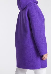 Lila fleecejacka med huva, med en texturerad yta och långa ärmar, sedd från sidan på en person.