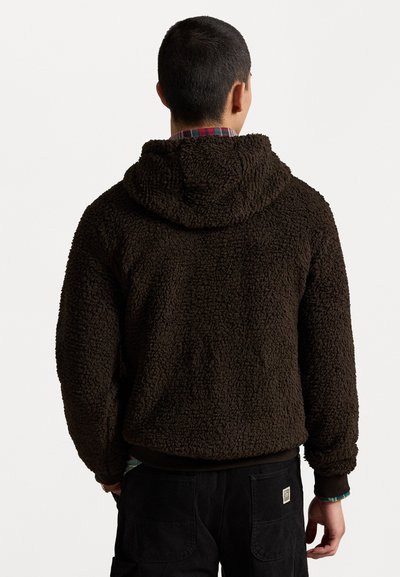 Bruine fleece hoodie met een grove, shaggy textuur en een capuchon, gedragen over een geruit shirt, gecombineerd met donkere broek.