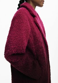 Flauschiger burgunderfarbener Mantel mit Reverskragen, seitlichen Taschen und strukturiertem Stoff. Das Design zeichnet sich durch ein weiches, dickes Aussehen aus.