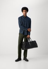 Kariertes Hemd in Marineblau und Grün, dunkle grüne Hose und eine große schwarze Lederdufflebag mit robusten Griffen.