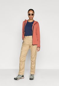 Κόκκινο φούτερ με φερμουάρ μπροστά, ναυτικό μπλουζάκι, μπεζ παντελόνι και γκρι ορειβατικά παπούτσια. Το μοντέλο φοράει επίσης γυαλιά ηλίου και στέκεται μπροστά σε λευκό φόντο.