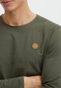 Long-sleeve olive green t-shirt in a soft fabric, featuring a circular tan patch logo on the chest with text and design details.