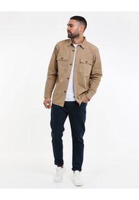 Beige overhemd met knoopsluiting en twee borstzakken, gecombineerd met donkerblauwe broek en witte sneakers. Man staat casual met een hand in zijn zak.