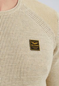 Beżowy sweter dzianinowy o teksturowanym wzorze, z czarną i złotą metką, na której widnieje napis "PME LEGEND". Okrągły dekolt i rękawy raglanowe.
