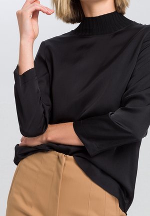 Schwarzes, seidiges Langarmoberteil mit einem gerippten, hohen Kragen, kombiniert mit hellbraunen, maßgeschneiderten Hosen. Details umfassen hochgekrempelte Ärmel und eine glatte Textur.