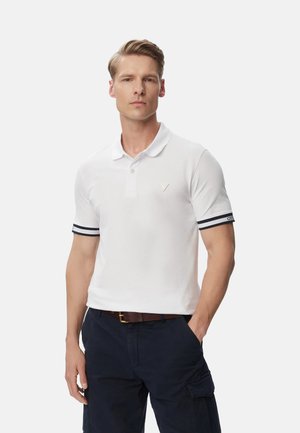 Uomo con capelli chiari che indossa una polo bianca con maniche a righe nere e pantaloni cargo blu navy, in piedi con una mano in tasca.