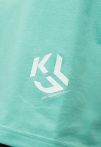 Τυρκουάζ ύφασμα με λευκό λογότυπο "KL" και το κείμενο "KARL LAGERFELD JEANS" τυπωμένο κοντά στο κάτω άκρο.