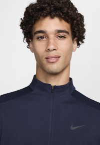 Chaqueta atlética azul marino con cuello de cremallera, hecha de tejido texturizado, que presenta un pequeño logo de Nike en el lado izquierdo del pecho y paneles de hombros en color contrastante.