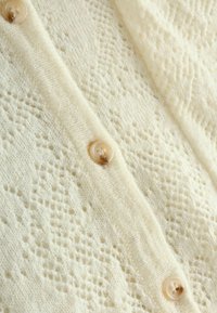 Cardigan lavorato a maglia color crema, caratterizzato da un motivo testurizzato e piccoli bottoni rotondi beige lungo la pattina anteriore. Tessuto morbido e leggero.