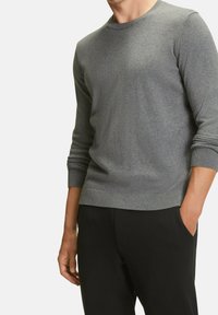 Grå stickad tröja med lång ärm, rundad halsringning, ribbade mudd och nederkant, bärs över svarta byxor. Slät textur med en avslappnad passform.