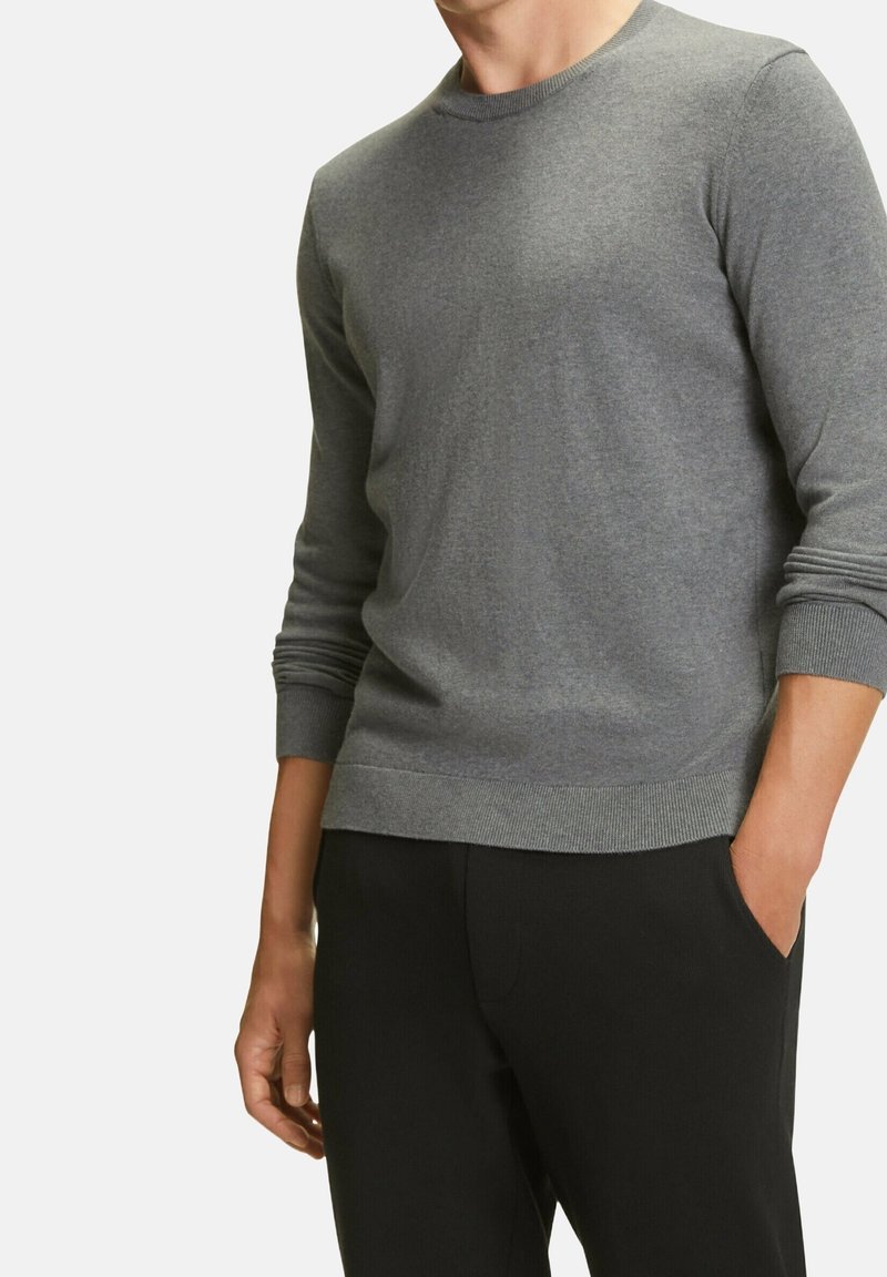 Grå stickad tröja med lång ärm, rundad halsringning, ribbade mudd och nederkant, bärs över svarta byxor. Slät textur med en avslappnad passform.