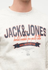 Κοντινό πλάνο σε ανοιχτό γκρι φούτερ με τη φράση "JACK & JONES" σε μεγάλους μπλε σκούρους και πορτοκαλί χαρακτήρες και μικρότερο κείμενο σχετικά με ποιοτικό βαμβάκι και νέα αισθητική.