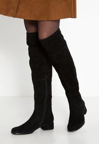 Botas de ante negras hasta la rodilla con tacón plano, que presentan una cremallera lateral y una textura suave, complementadas por un diseño ajustado.