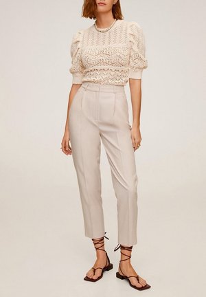 Vrouw die een crèmekleurige gehaakte blouse met pofmouwen draagt, beige broeken met hoge taille en zwarte vetersandalen met vierkante neus, staand tegen een effen achtergrond.