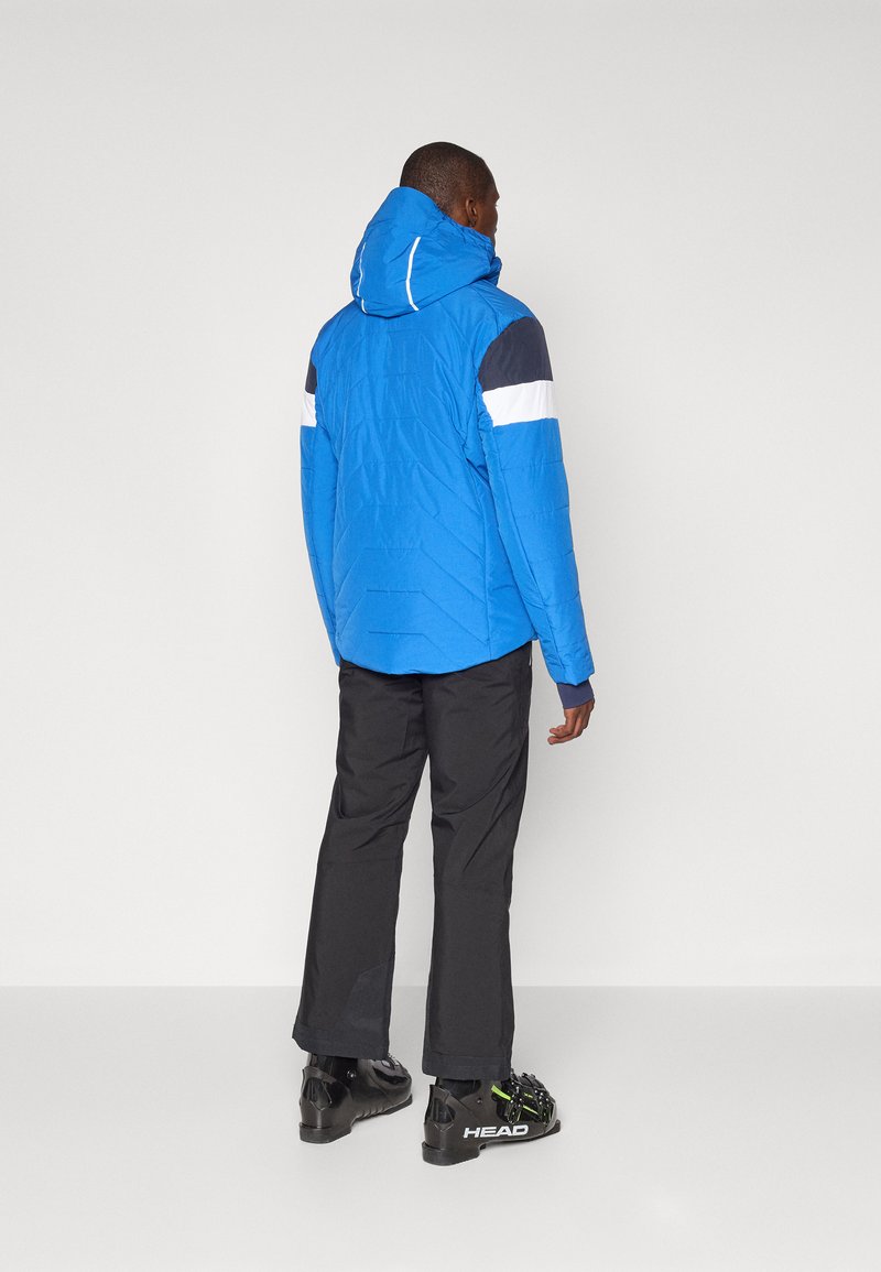 Blauwe pufferjack met capuchon, zwart-witte accenten. Gecombineerd met zwarte waterdichte broeken en zwarte skibotten met groene details.