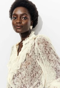 Blouse en dentelle ivoire avec un col à volants et un motif floral. La modèle porte des boucles d'oreilles en forme de cœur. L'arrière-plan est blanc.