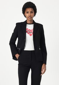 Černý blazer se dvěma knoflíky a kapsami na zip, nošený přes bílé tričko s červeným srdcem, spárovaný s černými kalhotami.